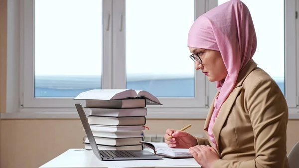 Wanita Muslim menulis dengan pena di notebook melihat ke dalam laptop — Stok Foto