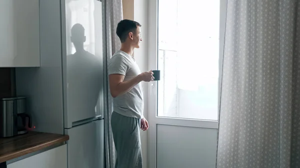 Guapo joven vierte café de la cafetera y se acerca a la ventana en la cocina — Foto de Stock