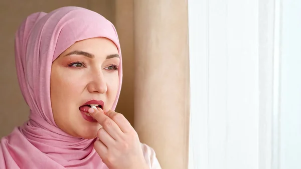 Ung kvinna i hijab dricker vit kapsel med vatten när hon står vid fönstret — Stockfoto