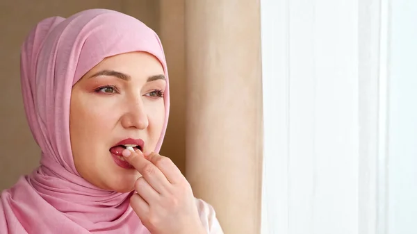 Mujer joven en hiyab bebe cápsula blanca con agua mientras está de pie junto a la ventana — Foto de Stock