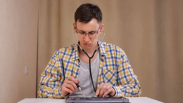 Человек в очках исследует разобранный ноутбук и слушает с помощью фонендоскопа — стоковое фото
