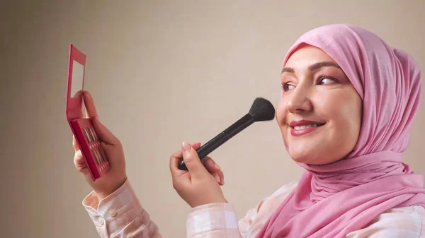 Mujer en hijab aplicando maquillaje con un cepillo mientras se mira en el espejo — Foto de Stock