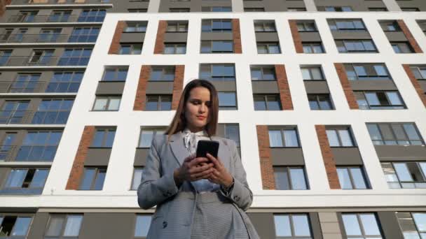 Mujer en un traje de negocios mira el teléfono y sonríe en el fondo de un edificio en un día soleado — Vídeos de Stock