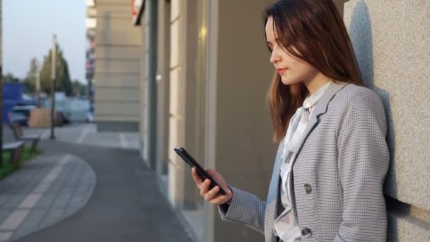 Giovane donna in giacca e cravatta guarda il telefono sulla strada vicino all'edificio — Video Stock