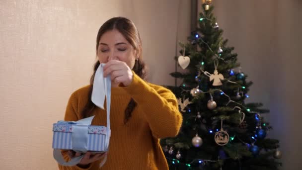 Vrouw in een mosterd trui maakt een lint van een geschenkdoos tegen de achtergrond van een versierde boom — Stockvideo