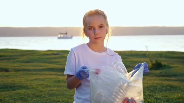 Adolescente mirando a la cámara sosteniendo una bolsa de basura recogida en la orilla del río — Vídeos de Stock