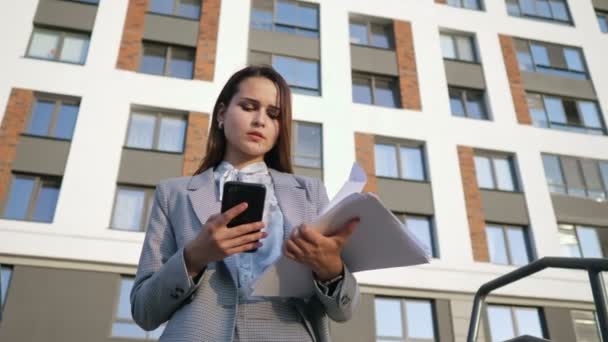 Mujer joven en un traje de negocios mira el teléfono y los documentos en el fondo del edificio, cámara lenta — Vídeos de Stock