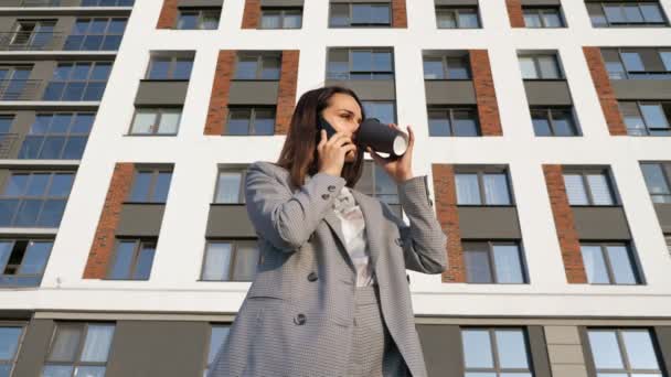 Женщина в деловом костюме пьет кофе из пластиковой чашки и разговаривает по телефону на фоне здания в солнечный день — стоковое видео