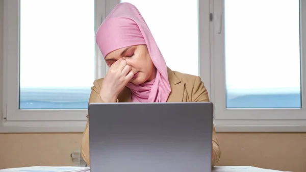 Kvinna i en hijab håller huvudet och masserar tempel medan du arbetar på en bärbar dator — Stockfoto