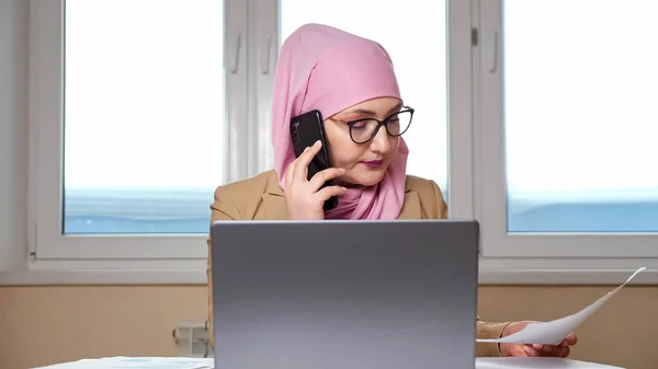 Γυναίκα σε μαντίλα εξετάζει έγγραφα και λειτουργεί σε φορητό υπολογιστή, ενώ μιλάμε στο τηλέφωνο — Φωτογραφία Αρχείου