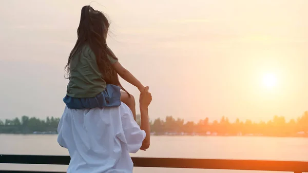 Mamá sostiene a su hija sobre los hombros mirando el río tranquilo — Foto de Stock