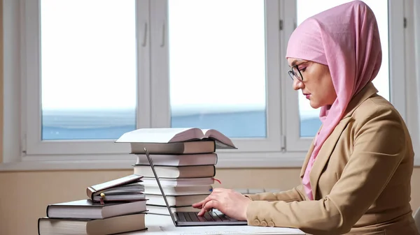 在一堆堆书籍中，一个穿着粉色头巾在笔记本电脑上打字的年轻穆斯林妇女的侧影 — 图库照片