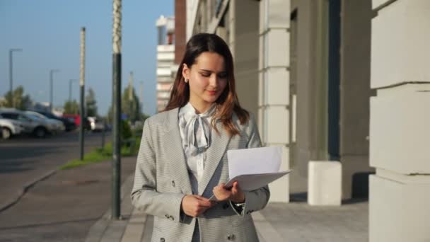 Mladá žena v obchodním obleku se dívá na dokumenty kráčející po ulici — Stock video