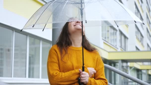 Eine glückliche junge Frau steht unter einem durchsichtigen Regenschirm im Regen vor dem Hintergrund eines Mehrfamilienhauses — Stockvideo