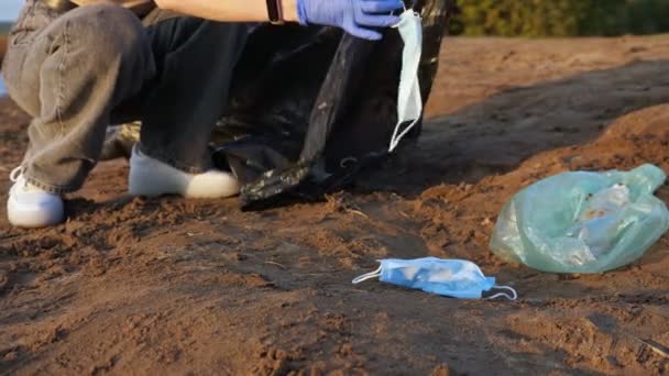 Nierozpoznawalna kobieta w rękawiczkach zbiera śmieci w czarnej torbie na brzegu rzeki, używane maski medyczne, zwolnione tempo — Wideo stockowe