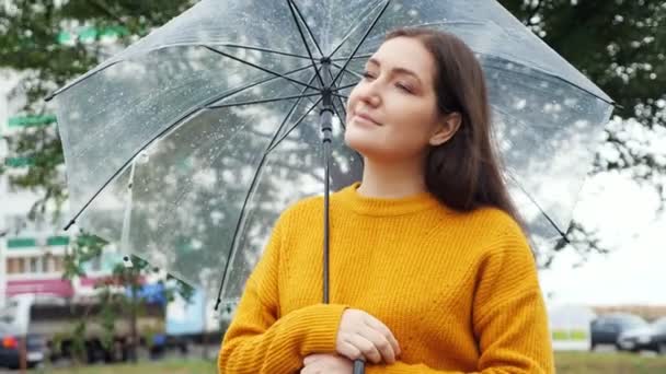 Jovem mulher em uma camisola de mostarda fica sob um guarda-chuva transparente em tempo chuvoso — Vídeo de Stock