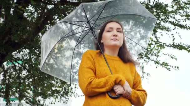 Trieste vrouw in een mosterdtrui staat en bevriest onder een transparante paraplu bij regenachtig weer — Stockvideo