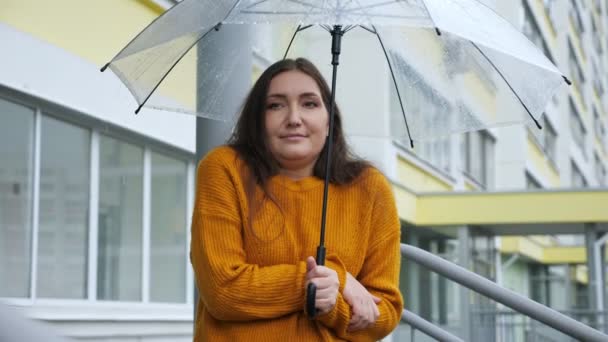 Молодая женщина стоит под прозрачным зонтиком под дождем в холодную погоду, замедленное движение — стоковое видео