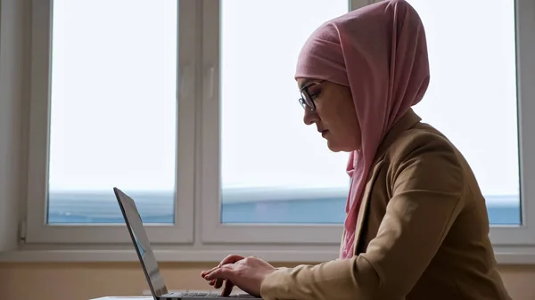 Γυναίκα σε ροζ χιτζάμπ δακτυλογράφηση σε φορητό υπολογιστή με φόντο παράθυρο — Φωτογραφία Αρχείου