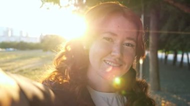 Kıvırcık kızıl saçlı kadın parkta selfie çekiyor.