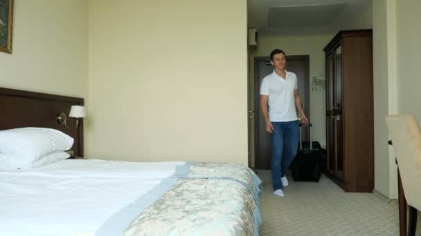 Young man with a suitcase enters the hotel room and falls contentedly on the bed — Stock Video