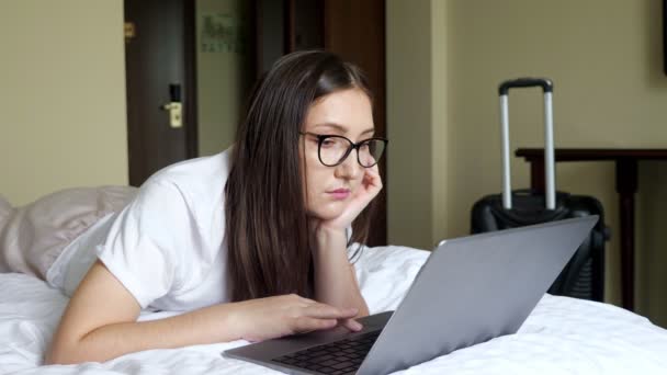 Young woman in glasses looks at a laptop lying on the bed on stomach against the background of a suitcase — Stock Video