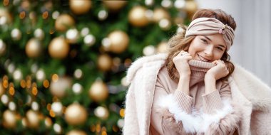 Sokakta süslenmiş köknar ağacının arka planında poz veren güzel, mutlu bir kızın açık hava portresi. Şık, kalın giysiler giyen bir manken. Noel, yeni yıl, kış tatili konsepti. Şehir yaşam tarzı.