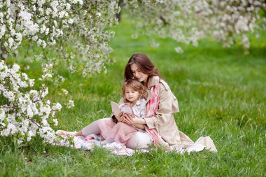 Anne ve küçük kızı birbirlerine sarılıyorlar ve elma bahçesinde yürüyorlar. Bahar hikayesi. Güzel bir bahar gününde mutlu bir aile. Bahar bahçesinde kitabı olan mutlu bir anne ve kızı.