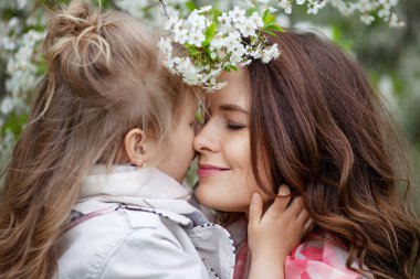 Anne ve küçük kızı birbirlerine sarılıyorlar ve elma bahçesinde yürüyorlar. Anne çocuğunu seviyor. Bahar hikayesi. Mutlu bir aile güzel bir bahar gününde