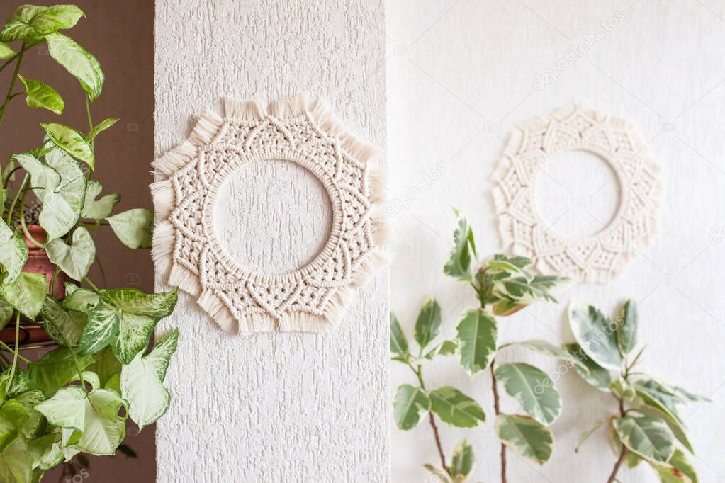 Cotton macrame mandala wall decoration hanging on white wall with green leaves. Handmade macrame wreath. Natural cotton thread. Eco home decor.