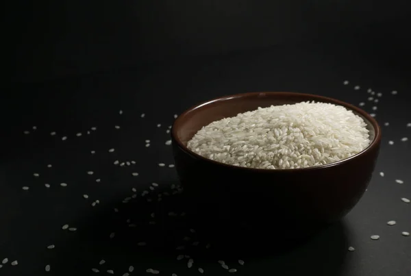 White uncooked rice in a bowl with scattered grains nearby on a black background
