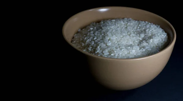 Riz blanc non cuit dans un bol avec des grains épars à proximité sur un fond noir — Photo