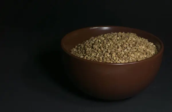 Brown uncooked buckwheat in a bowl with scattered grains nearby on a black background — Stock Photo, Image