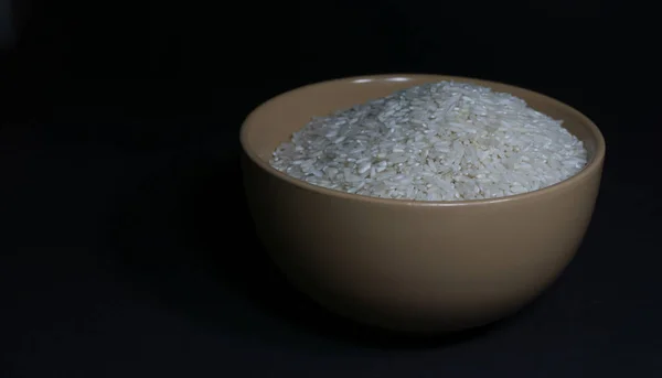 Riz blanc non cuit dans un bol avec des grains épars à proximité sur un fond noir — Photo
