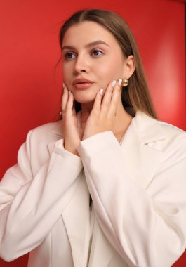 A young woman with long hair poses thoughtfully against a striking red background. She gently touches her face, showcasing a stylish look in a white jacket. The scene has a contemporary vibe.