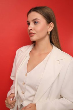 A woman stands confidently in a stylish white blazer and top, showcasing her elegance against a bright red background. The atmosphere is lively and fresh, highlighting her poised look.