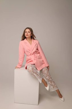 Bir model, pembe bir ceket ve dantel pantolon giyerek beyaz bir küpün üzerinde oturarak tarz sergiler. Tarafsız arka plan onun modaya uygun görünümünü arttırıyor, özgüvenini ve zarafetini gösteriyor..
