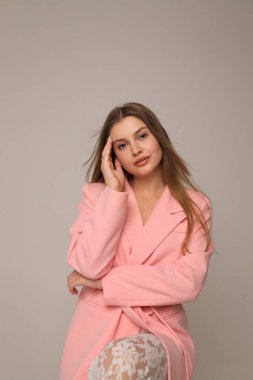 Genç bir kadın yumuşak pembe bir palto içinde duruyor, uzun saçları yavaşça dökülüyor. Yüzünde bir elle poz veriyor, minimalist bir ortamda düşünceli bir ifade sergiliyor..