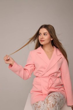 Genç bir kadın zarif bir şekilde stüdyoda oturuyor pembe bir ceket ve dantel tayt giyiyor. Uzun saçlarıyla oynuyor, rahat ama dengeli bir tavır sergiliyor..