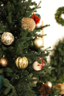 Noel ağacından sarkan renkli süsler, sıcak aydınlatılmış bir odada şenlikli bir atmosfer yaratıyor. Süslemeler, ışığı yakalayan altın ve kırmızı mücevherleri içeriyor..