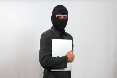 Siyah kar maskeli ve takım elbiseli bir adam şüpheli bir şekilde gümüş bir dizüstü bilgisayar tutuyor. Veri hırsızlığı, siber güvenlik tehdidi ve suç faaliyeti kavramı.