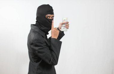 Takım elbiseli ve kar maskeli bir hırsız elinde bir tomar parayla sessizlik sinyali veriyor.