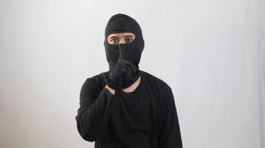 Siyah takım elbiseli ve siyah maskeli hacker beyaz arka planda izole edilmiş sessizlik işareti.