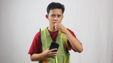 Yeşil yelek giyen Asyalı adam akıllı telefon tutuyor ve park görevlisi olarak ıslık çalıyor.