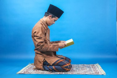 Müslüman bir adam, mavi arka planda bir paspasın üzerinde kitap okuyor.