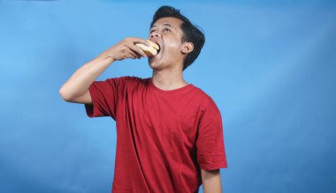 Kırmızı tişört giyen Asyalı bir adam mavi arka planda zevkle hamburger yiyor.