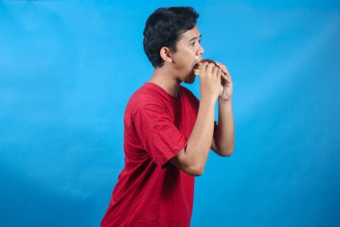 Kırmızı tişört giyen Asyalı adam mavi arka planda zevkle hamburger yiyor.