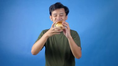Asyalı adam yeşil tişört giyip mavi arka planda zevkle hamburger yiyor.