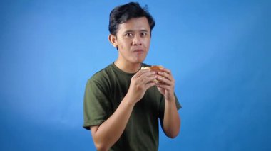 Asyalı adam yeşil tişört giyip mavi arka planda zevkle hamburger yiyor.