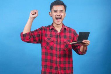 Asyalı adam, mavi arka planda izole edilmiş mutlu ifadeyle el kaldırırken açık tasarruf kitabını tutuyor.